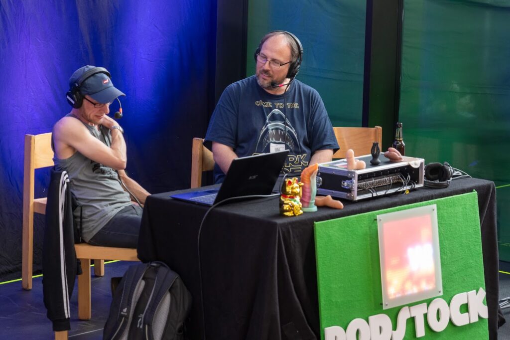 Toby und Jörn sitzen auf der Bühne. Am Tisch ist ein grünes Schild mit der Aufschrift "Podstock" angebracht. Beide tragen Headsets. Jörn spricht mit Toby, der seine rechte Hand auf die linke Schulter gelegt hat und nach unten schaut.