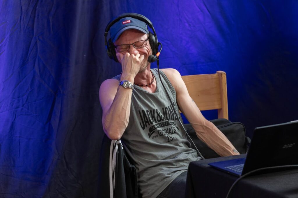 Toby sitzt auf der Bühne und trägt ein graues Tanktop, ein blaues Basecap und ein Headset. Er hat die Augen geschlossen und die Hand vor dem Mund und lacht.